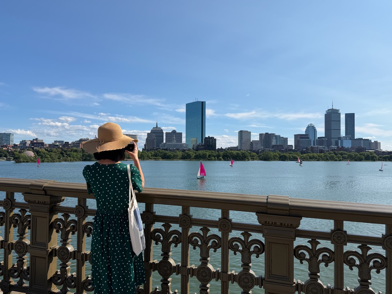 Me at the railing of the Longfellow Bridge, holding up a phone toward the Back Bay skyline across the Charles, on a sunny afternoon with sailboats on the water.